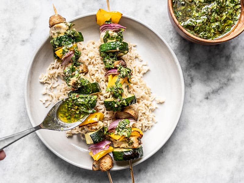 Chimichurri being drizzled over two chicken kebabs on a plate with a bed of rice.