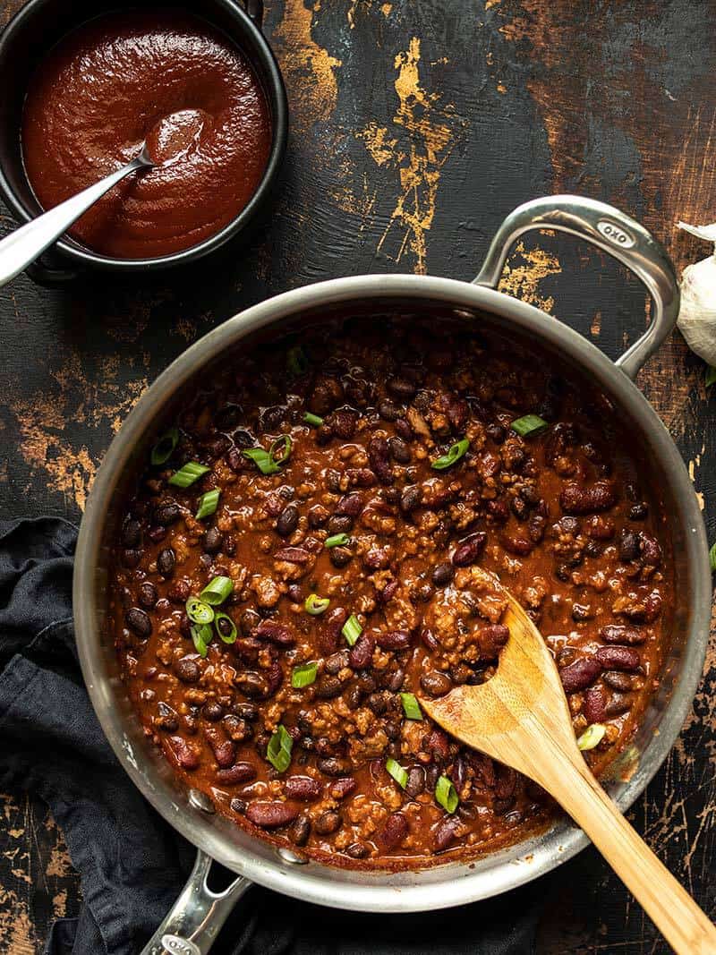 A skillet full of BBQ Beef and Beans with a wooden spoon and a black ceramic bowl of BBQ sauce on the side.