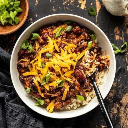 Overhead view of a bowl full of BBQ Beef and Beans, served with rice and topped with cheddar and green onion. A bowl of green onion on the side.