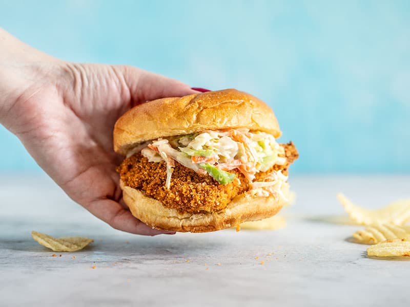 A hand holding a Baked Spicy Chicken Sandwich against a blue background.