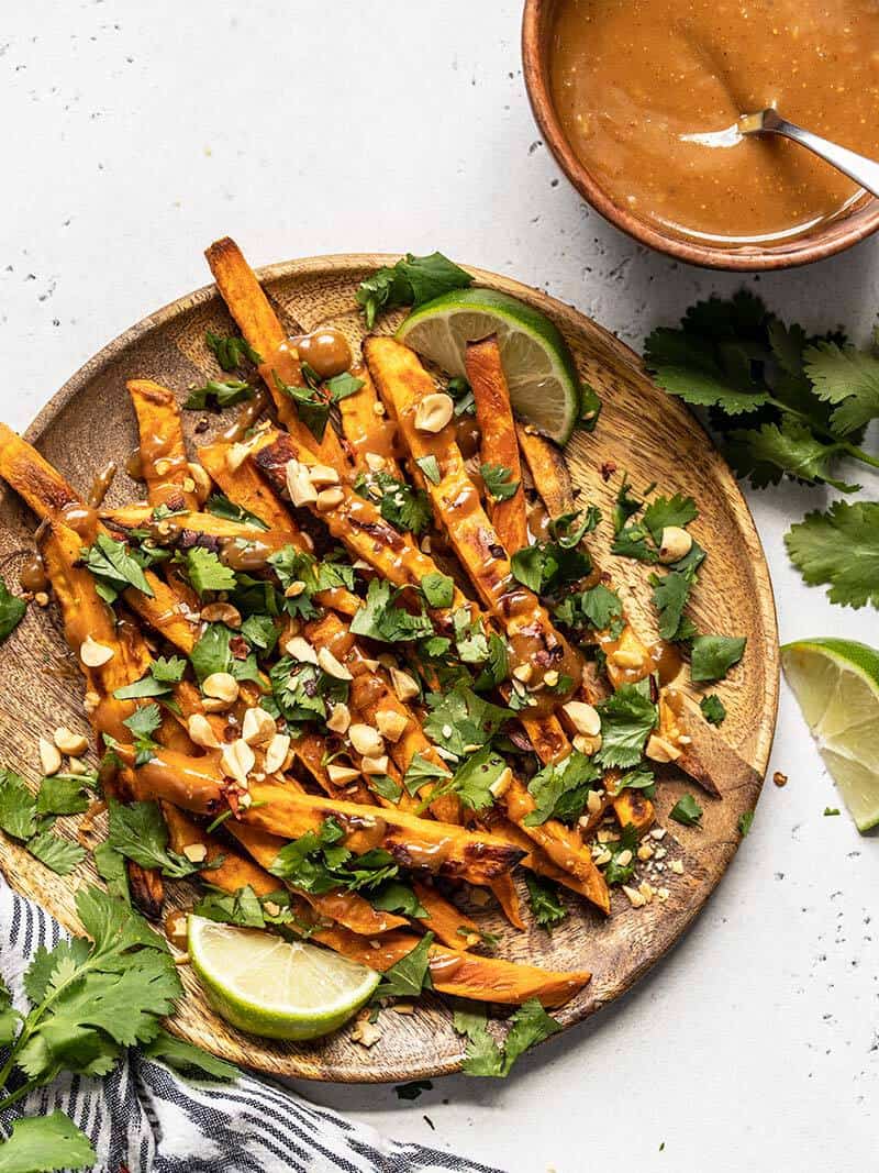 A wooden plate full of baked sweet potato fries with peanut lime dressing, cilantro, peanuts, lime wedges, and a bowl of dressing on the side.