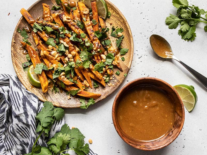 A plate of baked sweet potato fries loaded with cilantro and peanuts next to a bowl of peanut lime dressing.