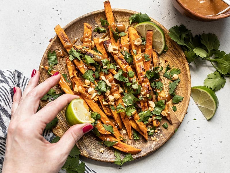 A wedge of lime being squeezed over a plate of baked sweet potato fries with peanut lime dressing.