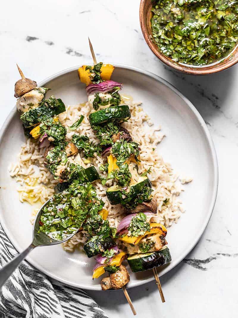 A spoonful of Chimichurri being drizzled over the Chimichurri Chicken Kebabs on a bed of rice.