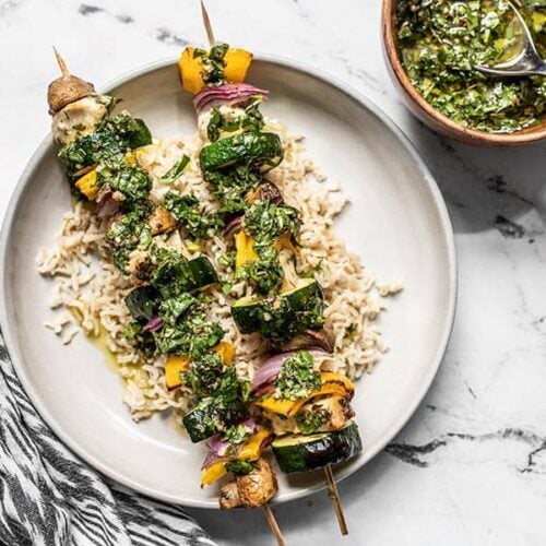 Two chimichurri chicken kebabs on a bed of brown rice, next to a bowl of chimichurri.