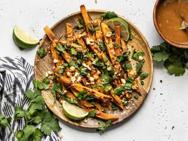 A wooden plate full of baked sweet potato fries drizzled with peanut lime dressing and topped with cilantro, peanuts, and red pepper.