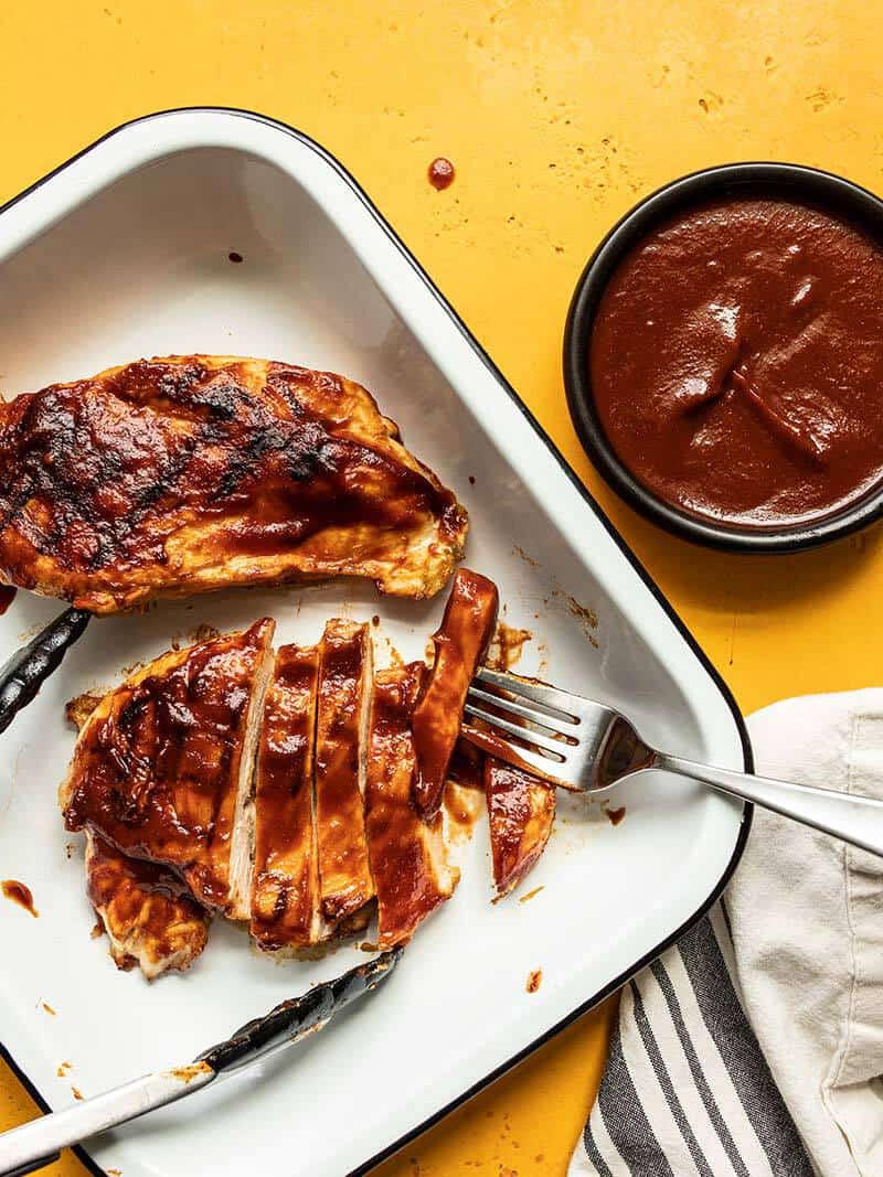 BBQ chicken breasts in a white dish, one sliced into strips, and a bowl of homemade bbq sauce on the side.