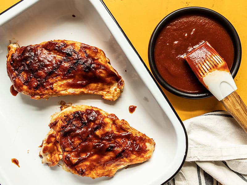 Two grilled chicken breasts coated in homemade bbq sauce with a dish of sauce and brush on the side.