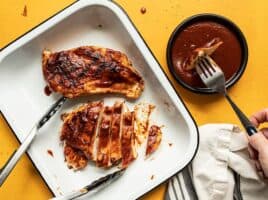 Two grilled chicken breasts coated in homemade bbq sauce in a dish, one sliced into strips, and a fork dipping a piece of chicken into a bowl of sauce.