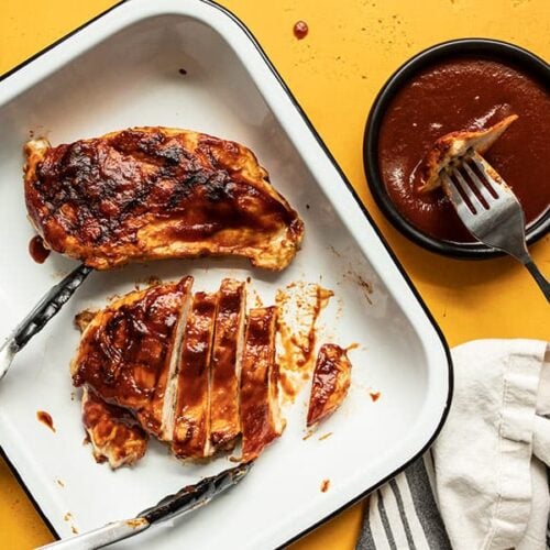 Two grilled chicken breasts coated in homemade bbq sauce in a dish, one sliced into strips, and a fork dipping a piece of chicken into a bowl of sauce.