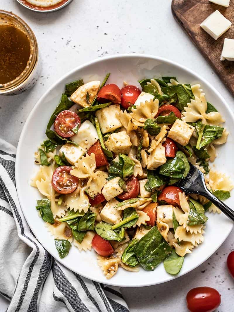 A big bowl of Tomato Mozzarella Pasta Salad with a jar of dressing and cubed mozzarella on the sides.