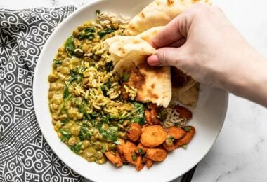 A hand dipping a piece of naan into the Creamy Coconut Curried Lentils with Spinach on a plate with curry roasted carrots