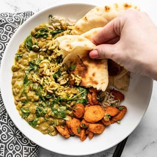 A hand dipping a piece of naan into the Creamy Coconut Curried Lentils with Spinach on a plate with curry roasted carrots