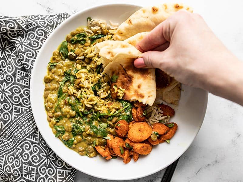 A hand dipping a piece of naan into the Creamy Coconut Curried Lentils with Spinach on a plate with curry roasted carrots