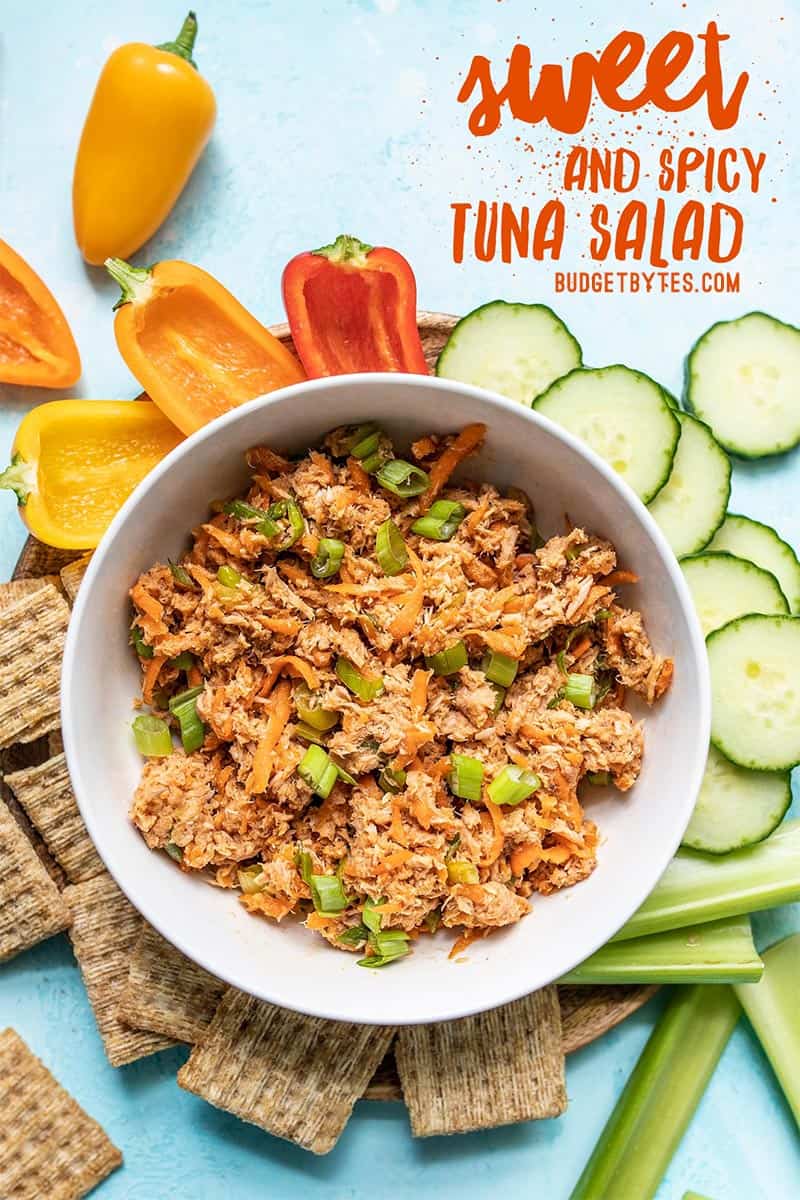 A bowl of Sweet and Spicy Tuna Salad surrounded by crackers and vegetables with title text overlay at the top.