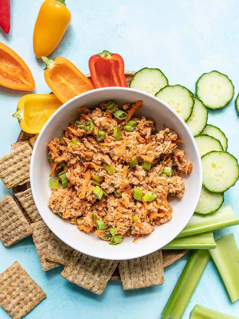 A bowl of Sweet and Spicy Tuna Salad surrounded by crackers and vegetables on a blue surface.