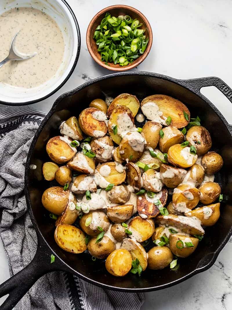 Overhead view of a cast iron skillet containing crispy potatoes with lemon dill tahini dressing, a bowl of dressing and bowl of green onion on the side