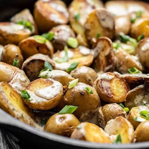Close side view of a skillet full of potatoes topped with tahini dressing and green onion