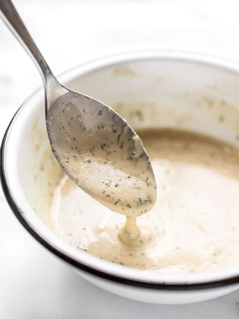 Lemon Dill Tahini Dressing dripping off a spoon into a white bowl, viewed from the side.