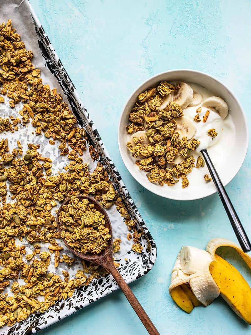 A sheet pan of Super Crunchy Oil Free Granola next to a bowl with yogurt, banana, and granola, and a half eaten banana.