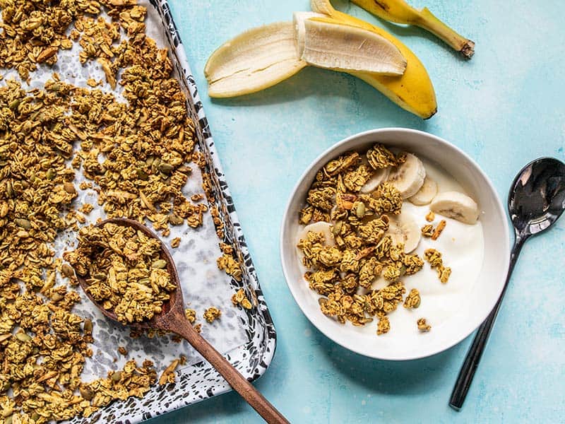 A sheet pan of Super Crunchy Oil Free Granola next to a bowl with yogurt and granola, and a half eaten banana.