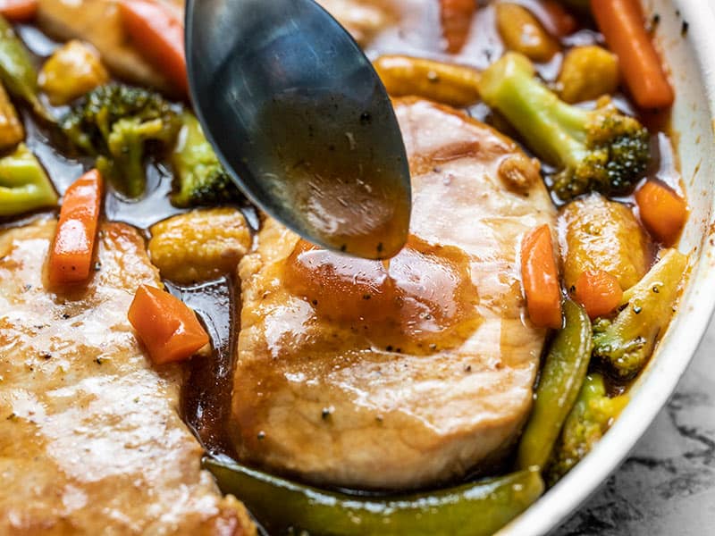 Close up of Sweet and Sour Sauce being spooned over a pork chop in the skillet, surrounded by vegetables and sauce.