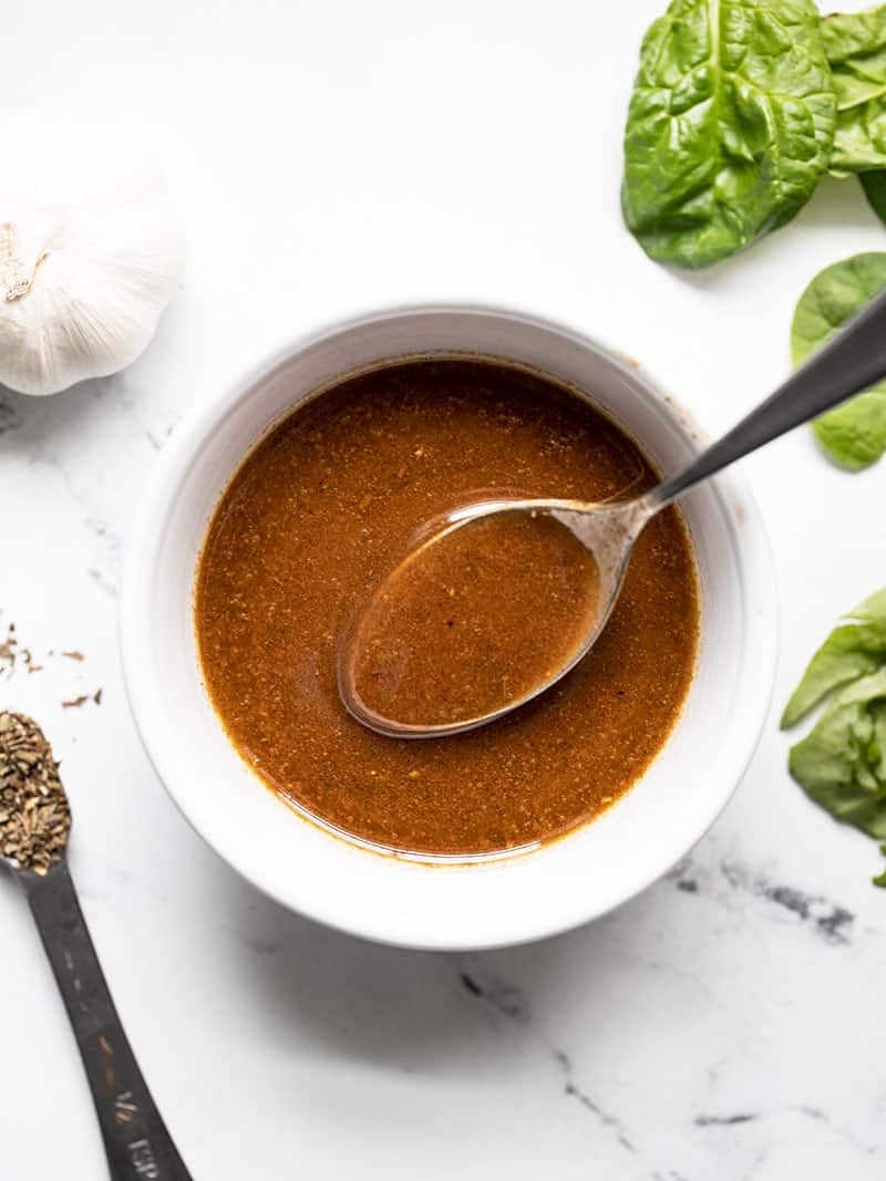 A small white bowl of homemade balsamic vinaigrette with a spoon in it and spinach, garlic, and dried basil on the side