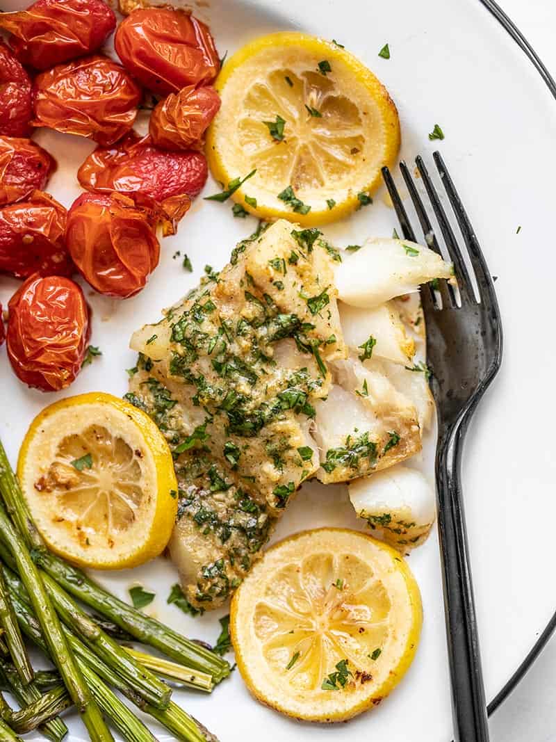 Garlic butter baked cod on a plate with tomatoes and asparagus, with a black fork on the side.