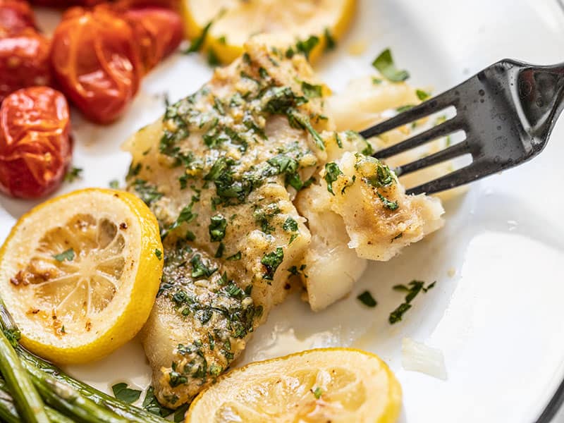 Close up of a fork digging into a filet of Garlic Butter Baked Cod on a plate with vegetables.