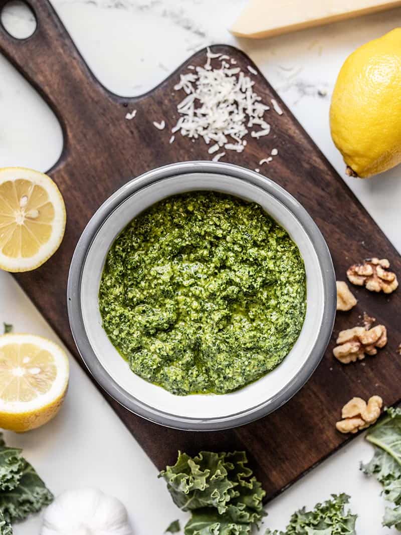 A bowl of Kale Pesto on a wooden cutting board surrounded by lemons, walnuts, kale, and Parmesan