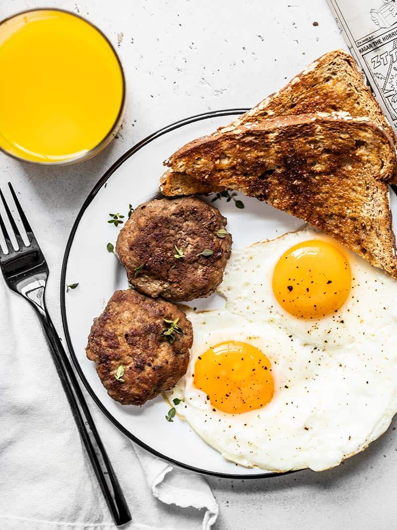 Two maple sage breakfast sausage patties on a plate with eggs and toast, and a glass of orange juice on the side.