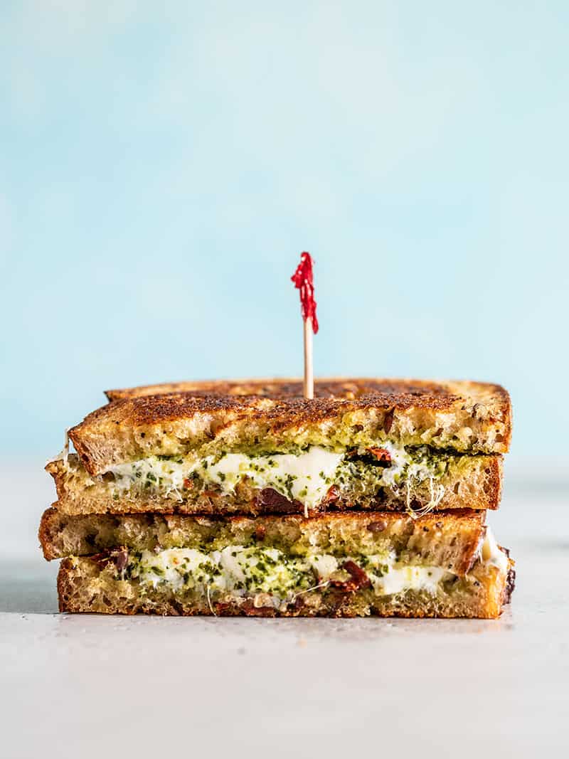 Two halves of a Pesto Grilled Cheese Sandwich stacked with cut sides facing the camera. Blue background.
