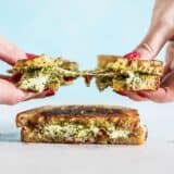 Two hands pulling the pesto grilled cheese sandwich in half, with the cheese pulling between and sun dried tomato pieces exposed.
