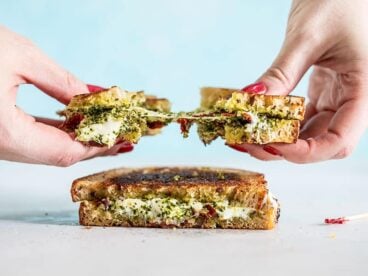 Two hands pulling the pesto grilled cheese sandwich in half, with the cheese pulling between and sun dried tomato pieces exposed.