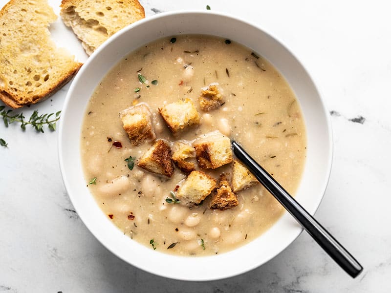 A bowl of Rosemary Garlic White Bean Soup with croutons and a black spoon in the middle