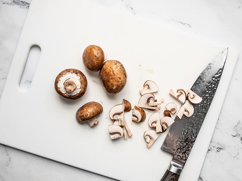 Baby bella mushrooms being sliced on a white cutting board