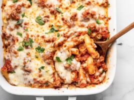 Overhead view of baked ziti being scooped out of the casserole dish with a wooden spoon.