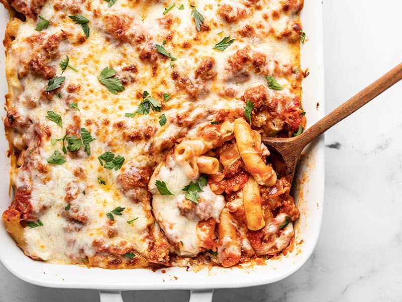 Close up of a wooden spoon in the casserole dish full of baked ziti