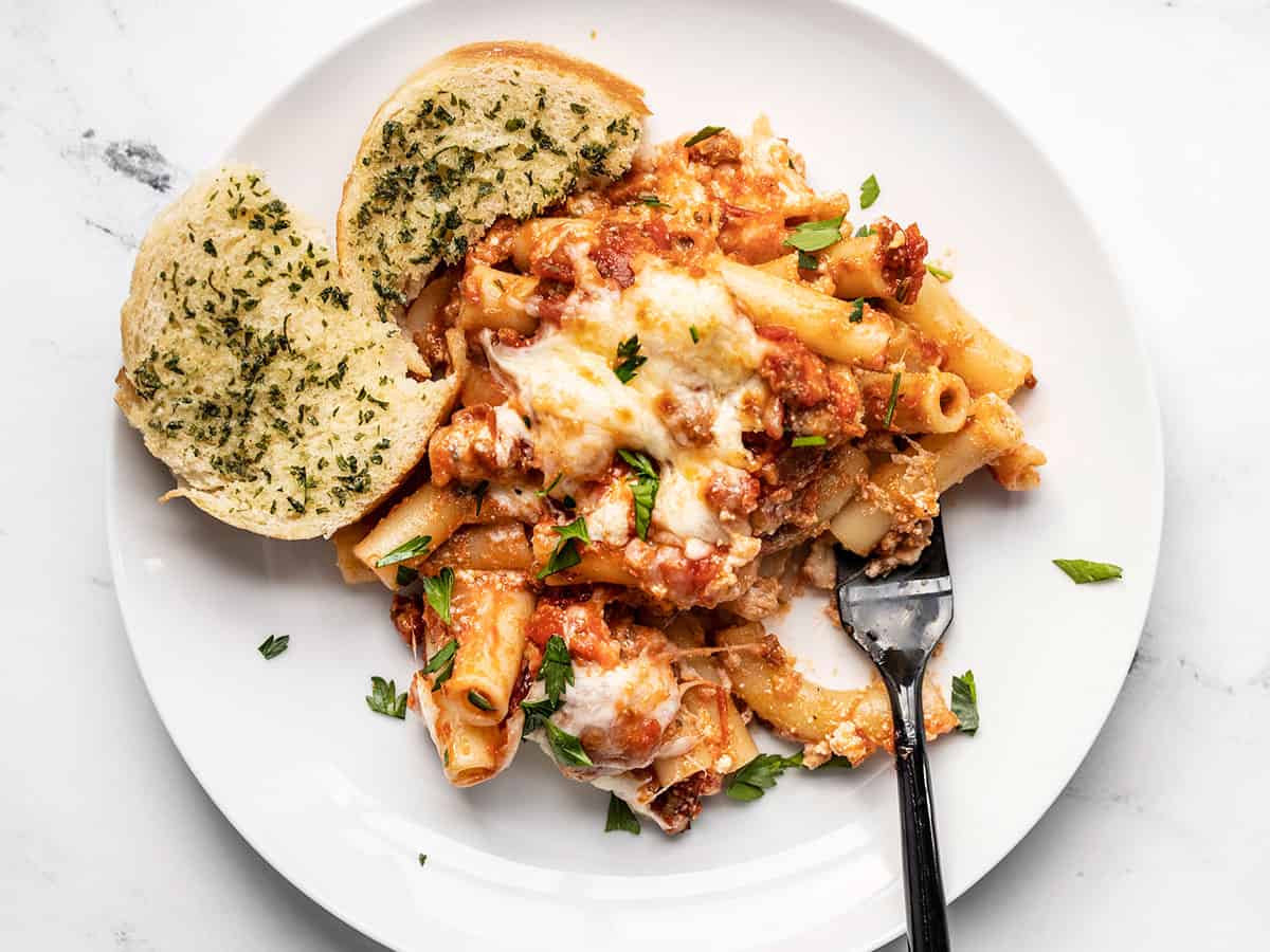 Baked ziti on a plate with garlic bread and a fork. 