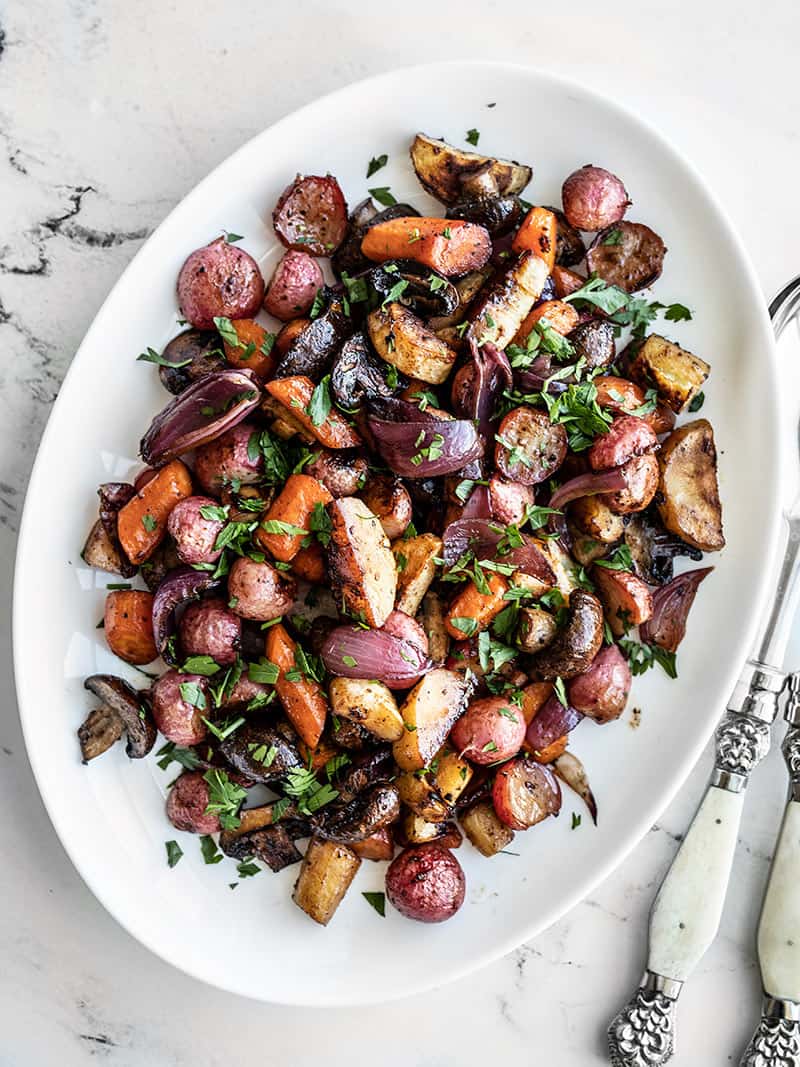 A white oval platter full of Balsamic Roasted Vegetables with metal serving ware on the side.