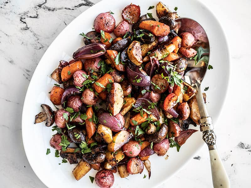 A serving spoon stuck in the side of a platter full of balsamic roasted vegetables, garnished with chopped parsley.