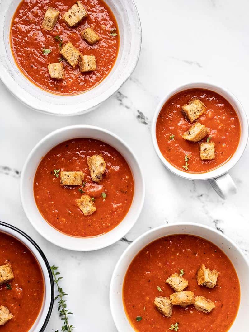Five different white bowls of Secret Ingredient Tomato Soup with whole grain croutons and fresh thyme garnishes