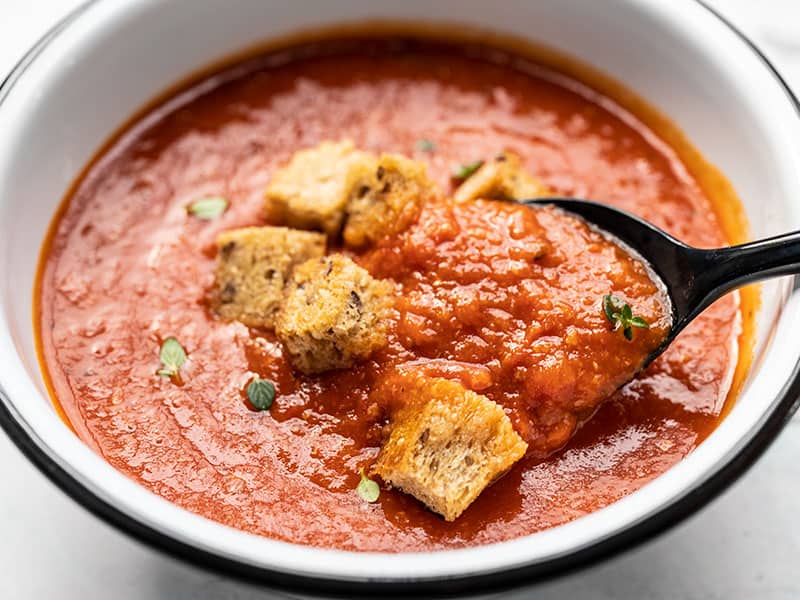 Close up of a bowl of secret ingredient tomato soup with whole grain croutons and a black spoon lifting a bite