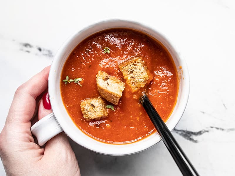 A hand holding a white mug full of secret ingredient tomato soup, garnished with croutons and thyme