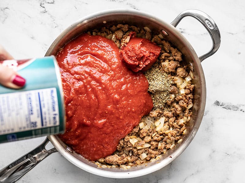 Crushed tomatoes being poured into the skillet, with sausage, onions, tomato paste, and Italian seasoning