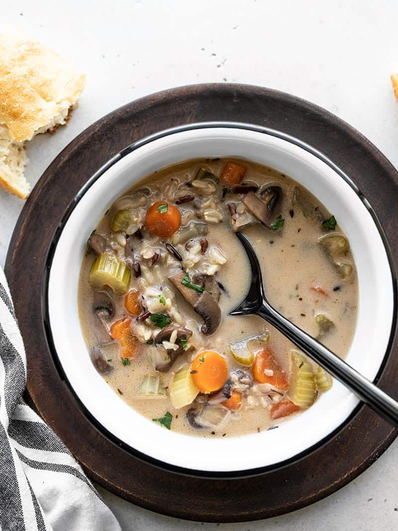 Overhead view of a bowl full of creamy vegetable wild rice soup on a wooden plate with a piece of bread on the side