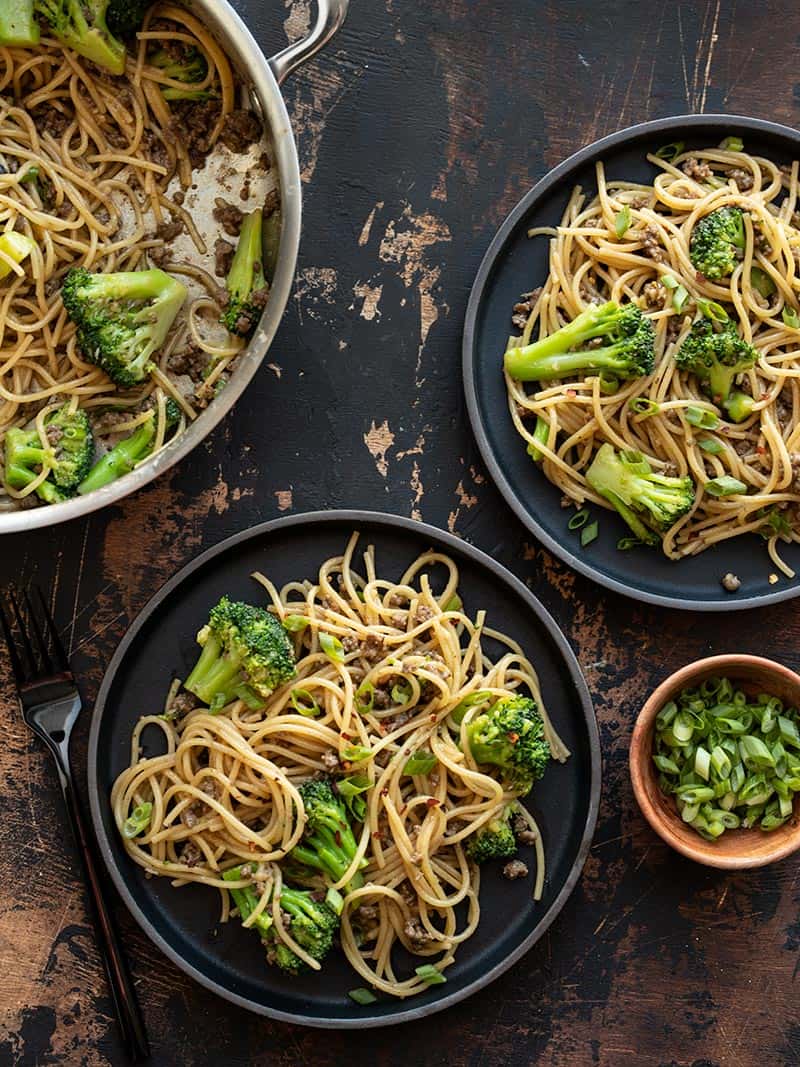 Two black plates of Garlic Noodles with Beef and broccoli next to the pan and a bowl of sliced green onions.