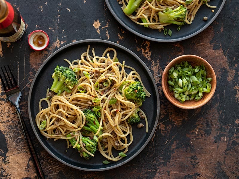 Two plates of Garlic Noodles with Beef and Broccoli next to a bottle of oyster sauce and a small bowl of sliced green onions