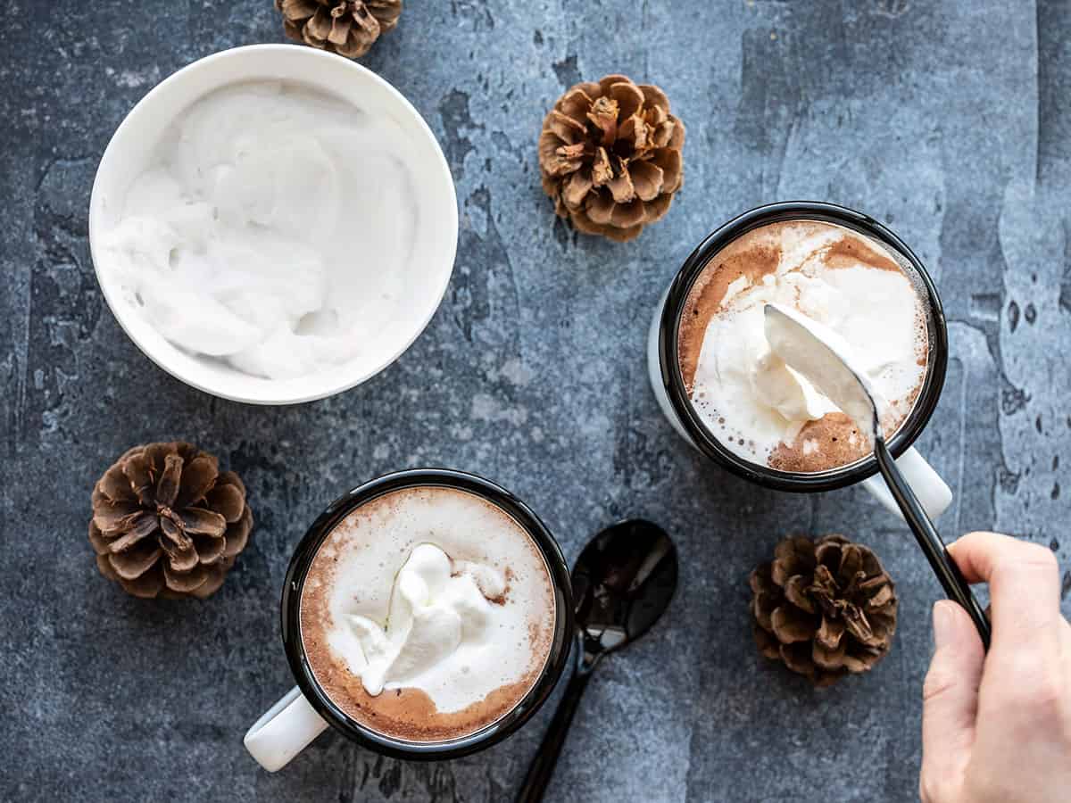 Overhead view of whipped cream being spooned onto two mugs of hot cocoa. 