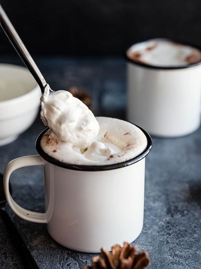 Homemade whipped cream being spooned onto a mug of hot cocoa, a second mug in the background